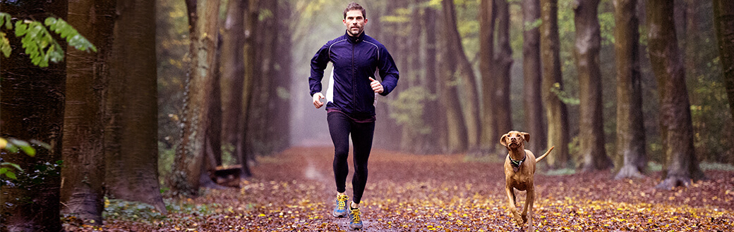 A fit man running outside on fallen leaves with a dog.